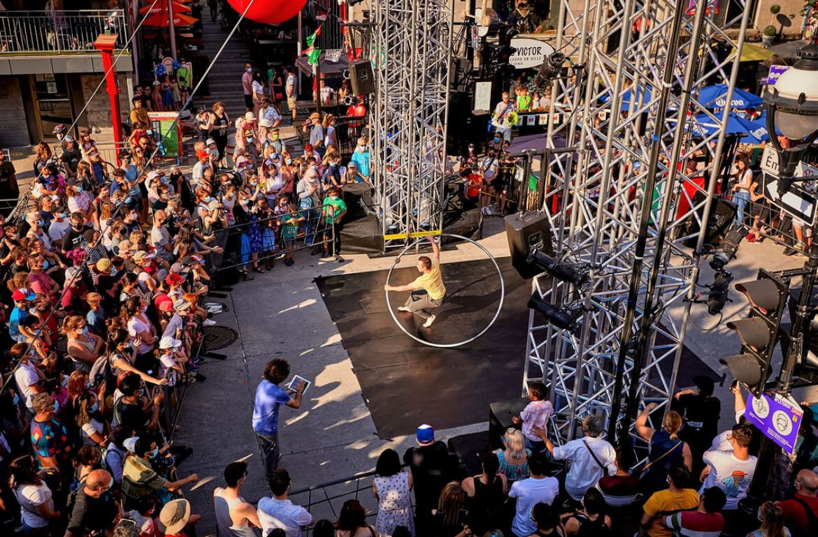 A crowd gathers at an outdoor festival to watch a performer with a large hoop on a sunlit stage, surrounded by urban infrastructure.