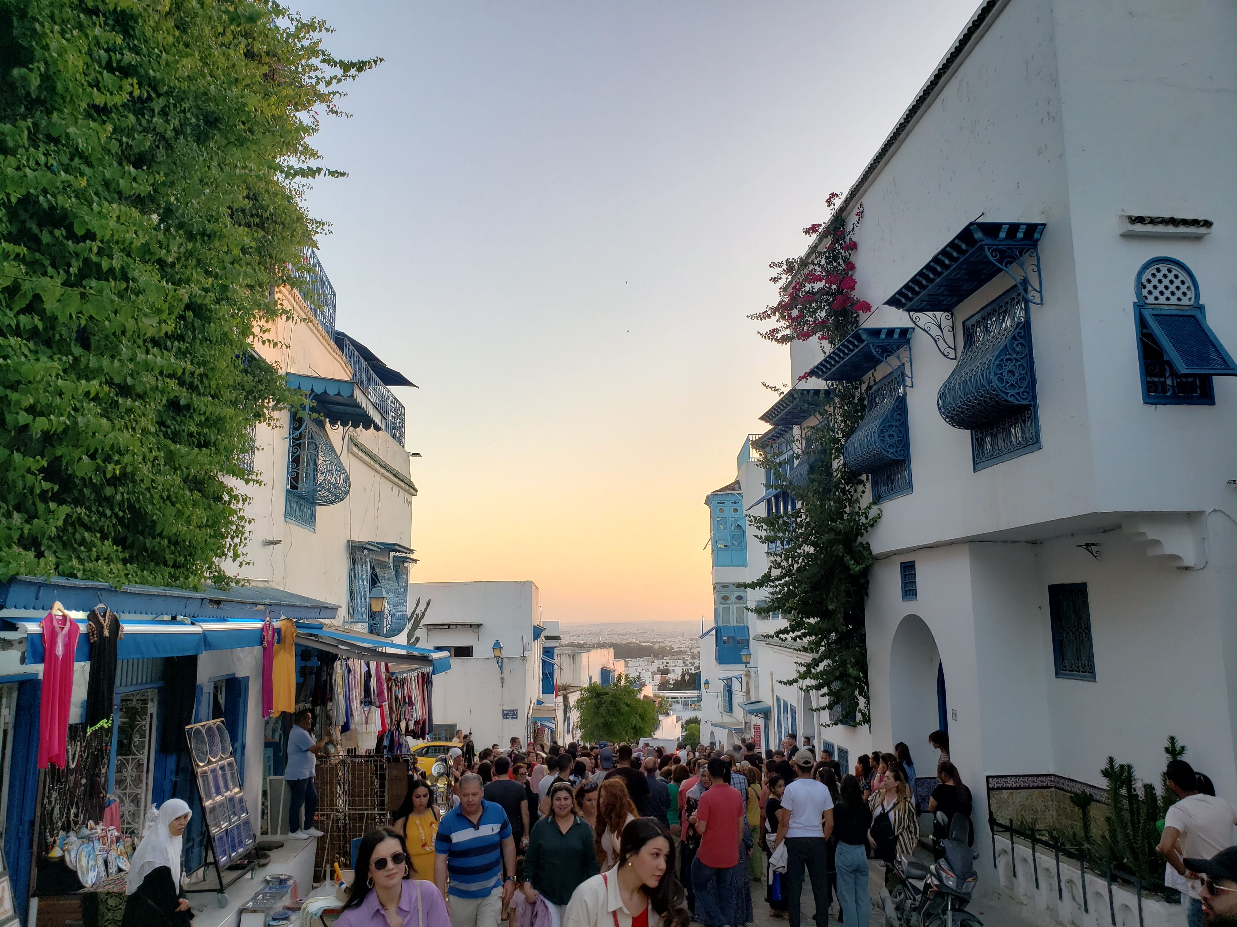 Crowded market street with traditional white and blue buildings at sunset, indicative of Mediterranean architecture.