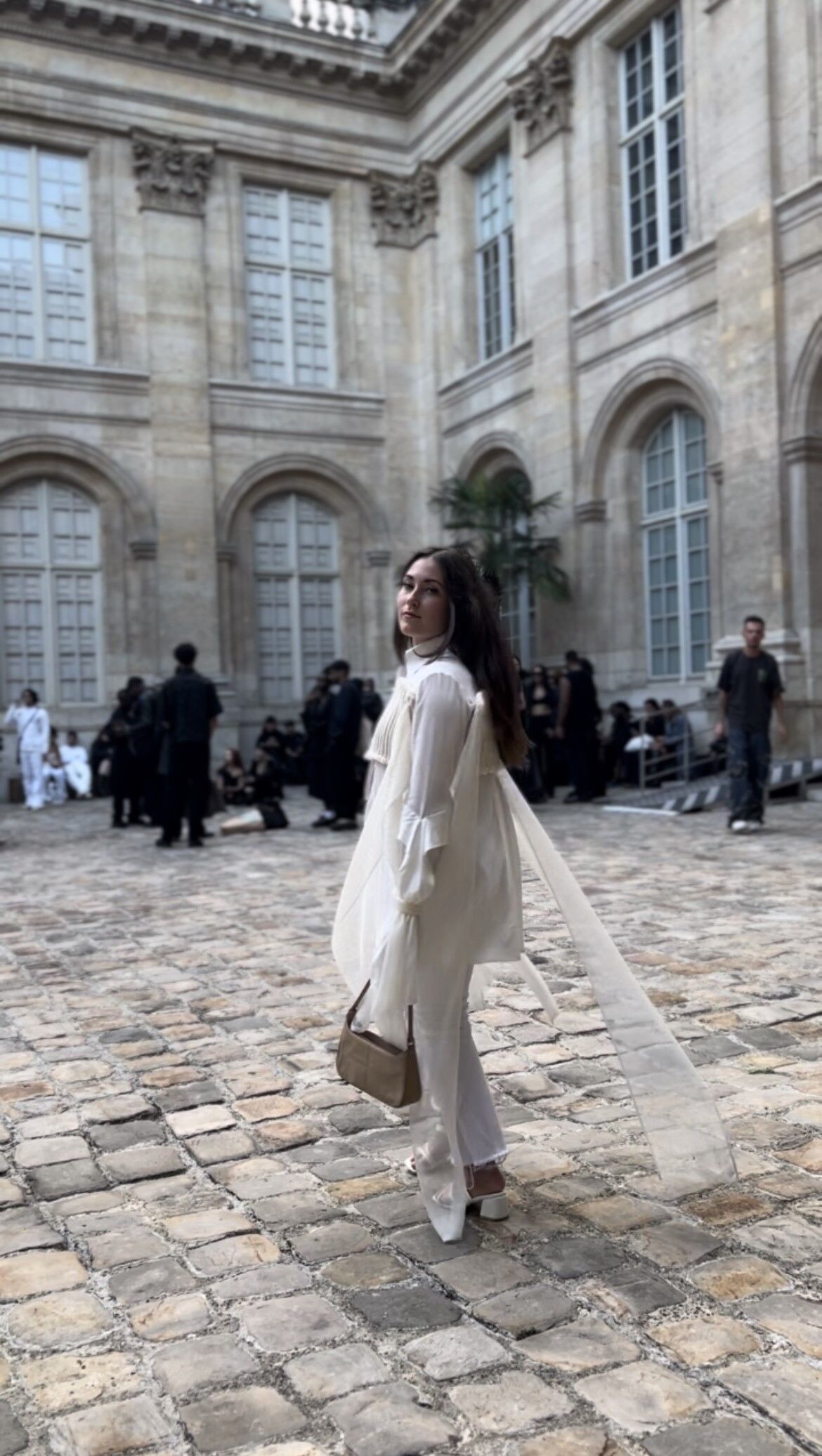 A woman in a white outfit posing at a fashion event, with classical architecture in the background.