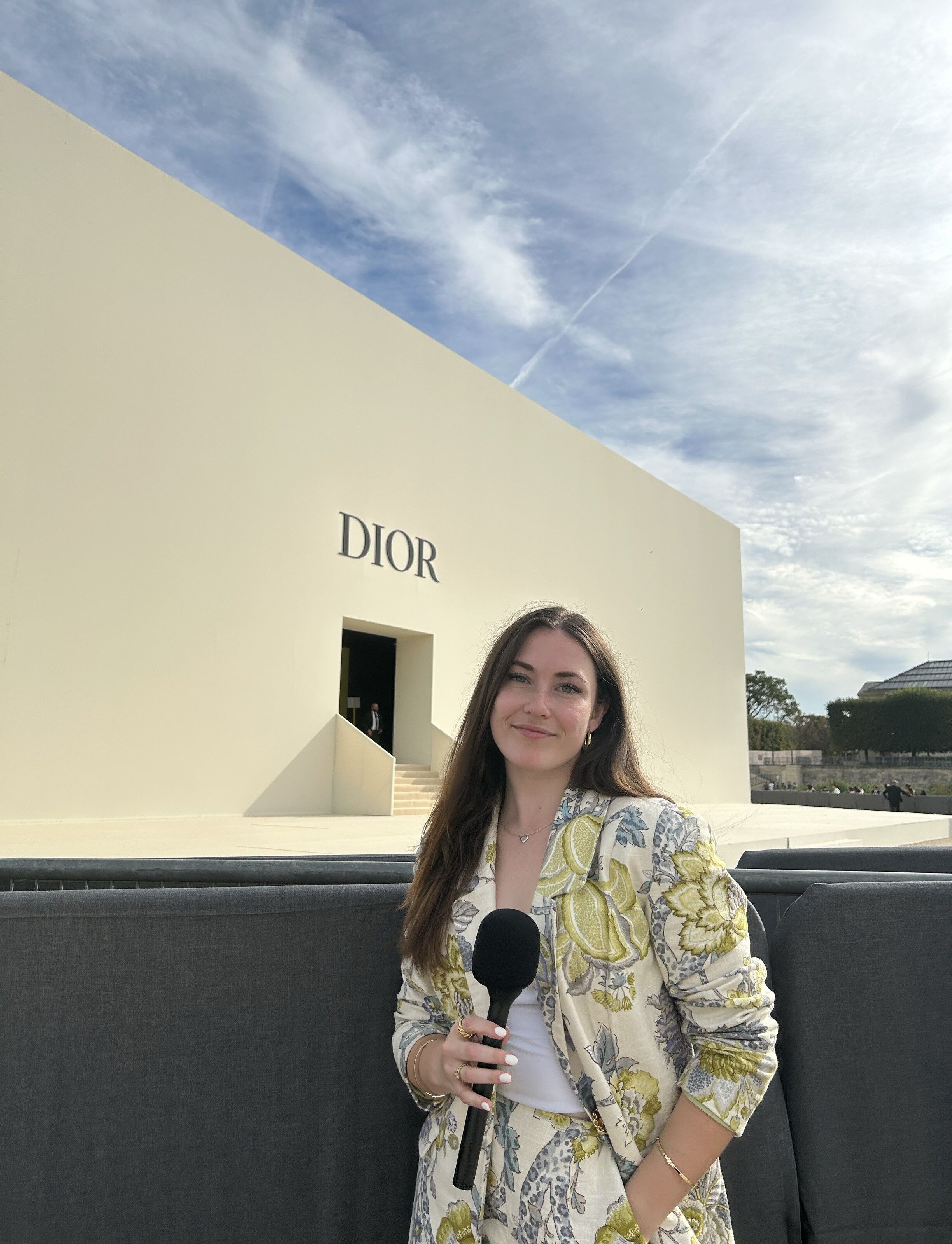 A reporter with a microphone stands before a Dior show venue, ready to broadcast.