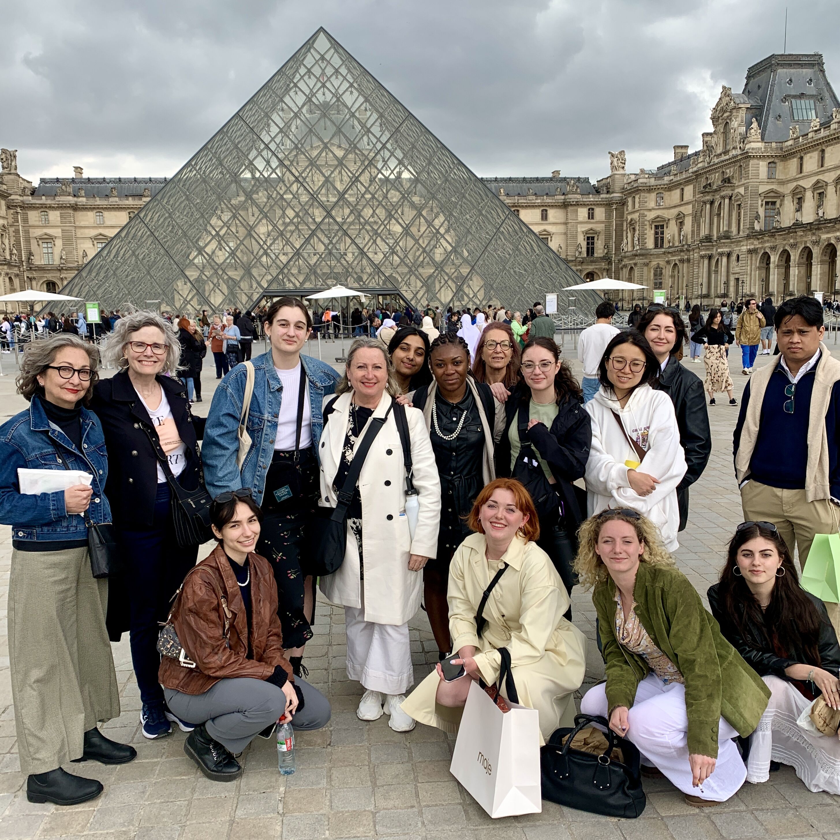 Un groupe multiculturel de douze touristes, affichant un mélange de tenues décontractées et élégantes, pose devant l'iconique pyramide du Louvre à Paris, capturant un moment de leur exploration culturelle.