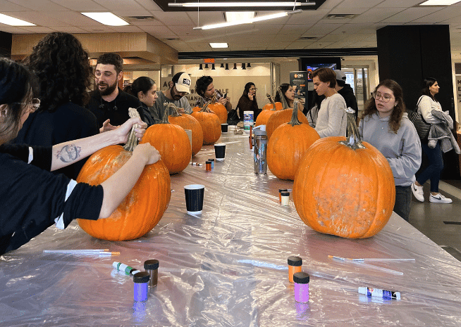 Students actively decorating pumpkins at a school event, surrounded by various art supplies on covered tables, creating a festive and engaging atmosphere.