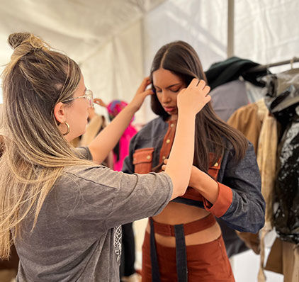  Une styliste concentrée assure l'ajustement d'une tenue en cuir sur un mannequin dans l'ambiance animée d'une tente de défilé de mode.