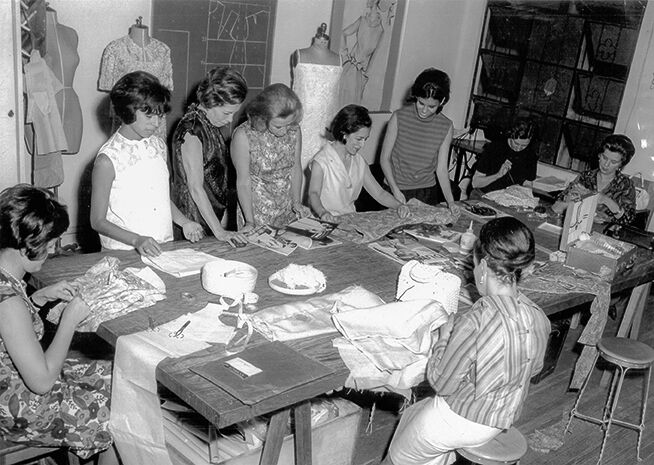 Fotografía en blanco y negro de ocho mujeres participando en actividades de costura alrededor de una mesa grande, con maniquíes y telas al fondo.