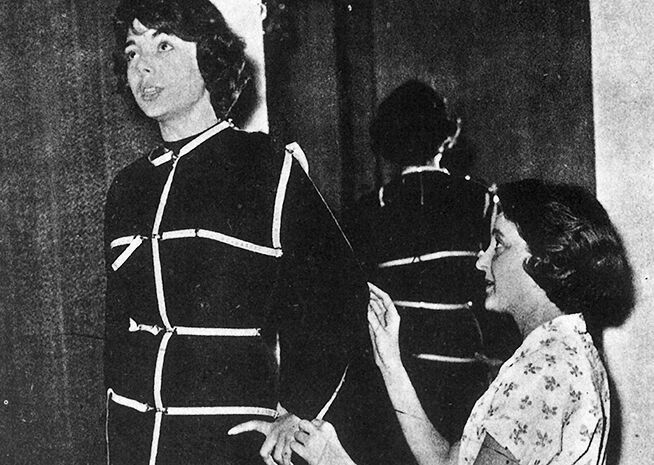 A seamstress measures a woman wearing a calibration suit against a mirror.