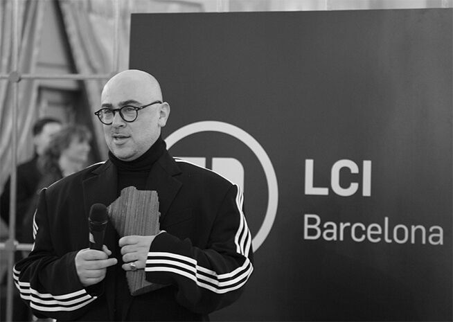 Un hombre calvo con gafas, vestido con cuello alto y chaqueta negra con rayas blancas en las mangas, sostiene un micrófono con fondo de LCI Barcelona.
