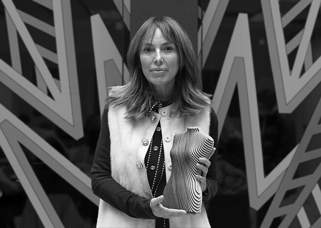 A woman holds an abstract sculpture award, dressed in a fur vest and polka dot blouse, with a geometric pattern backdrop.
