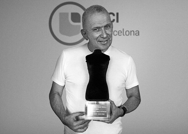 Un hombre sonriente con una camiseta blanca casual está de pie frente a un logotipo, sosteniendo un trofeo con una placa que probablemente lleva su nombre.