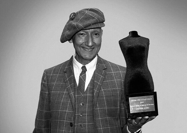 A cheerful man in a plaid suit and beret holds a sculpted award, radiating pride.