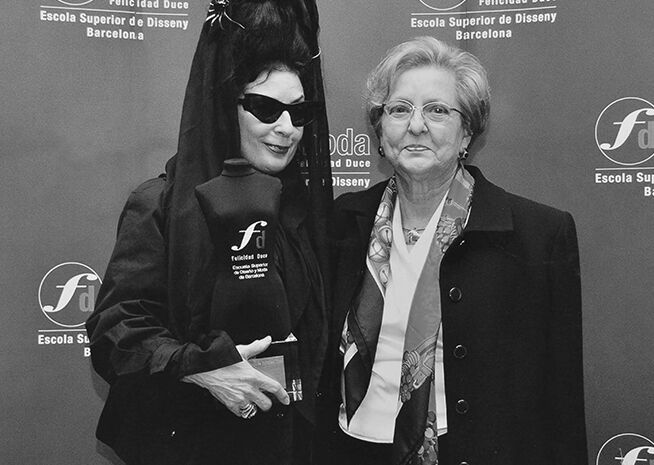 Dos mujeres posando en un evento de moda; una con atuendo vanguardista y gafas de sol, la otra con vestimenta académica formal.