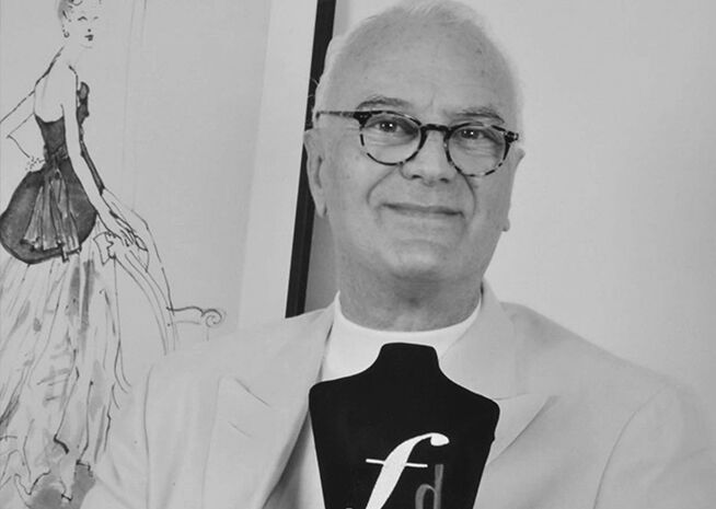 Foto en blanco y negro de un diseñador de moda sosteniendo un premio con un emblema 'f', con un boceto al fondo.