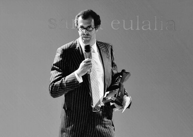 Foto en blanco y negro de un hombre con traje a rayas sosteniendo un micrófono y un premio, probablemente dirigiéndose a una audiencia.
