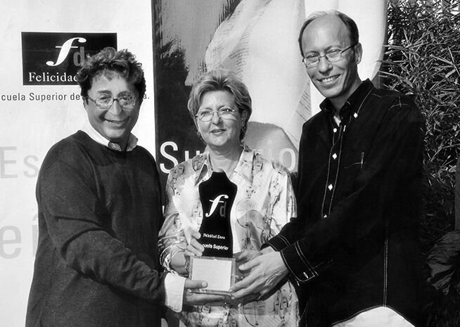 Tres individuos posan para una fotografía durante la entrega de un premio; una mujer en el centro sostiene un trofeo con inscripción, flanqueada por dos hombres sonrientes.