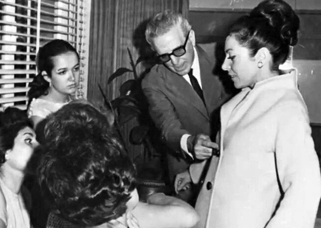 Black and white photo of an elderly gentleman in glasses, addressing a woman in a formal coat, surrounded by onlookers.