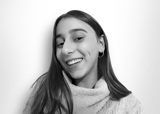 A youthful woman with a bright smile, donning a turtleneck and hoop earrings, is captured in a black and white photograph.