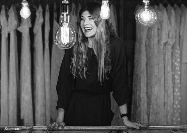 A woman in a black dress laughs heartily, surrounded by hanging filament light bulbs in a monochrome setting.