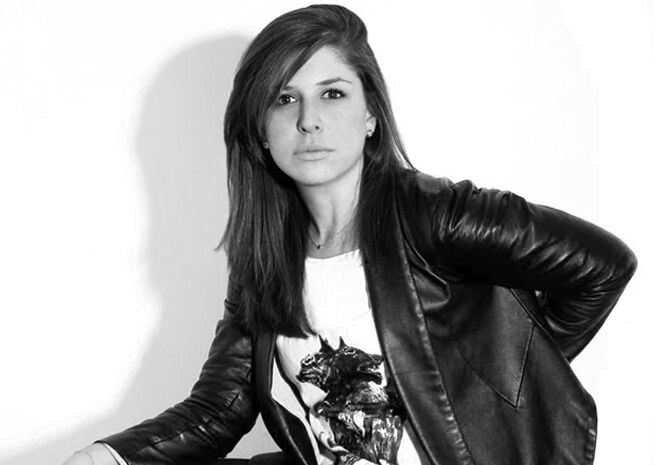 A poised young woman in a black leather jacket and graphic tee, exuding confidence against a plain background.