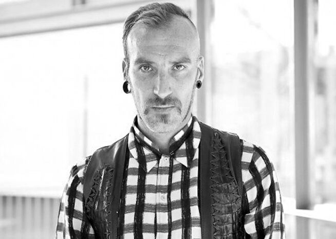 Black and white portrait of a man with a short haircut, mustache, ear gauges, and a patterned checked shirt. His direct gaze at the camera carries a neutral expression.