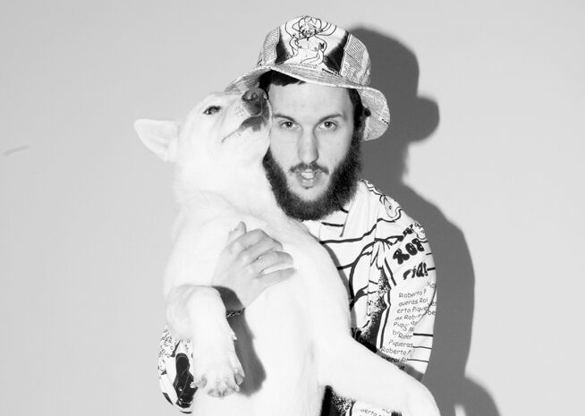 A black and white photograph of a bearded man wearing a printed shirt and bucket hat, embracing a large white dog with an open mouth.