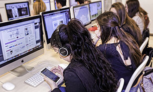 Estudiants concentrats en la seva feina en un laboratori d'informàtica, utilitzant ordinadors de sobretaula i dispositius personals amb auriculars.