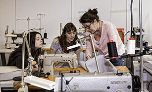 Tres mujeres participan en un taller de costura, usando una máquina de coser y enfocándose en la tela.