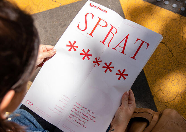 Una persona revisa un ejemplar tipográfico rojo con la palabra 'SPRAT' en un día soleado.