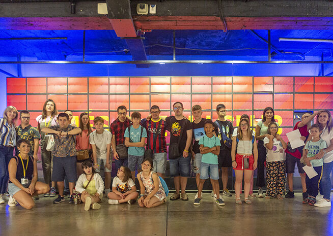 A diverse group of individuals of varying ages posing together during a visit to a vibrant, interactive exhibition space with colorful lighting in the background.