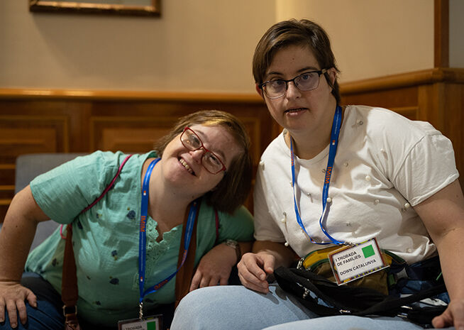 Una mujer alegre con gafas apoyándose en una amiga en una conferencia, ambas con lanyards, simbolizando camaradería.