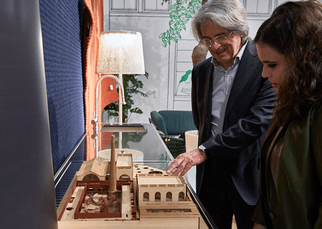 A man presents an architectural model to a viewer, pointing out details of the design within an indoor setting.
