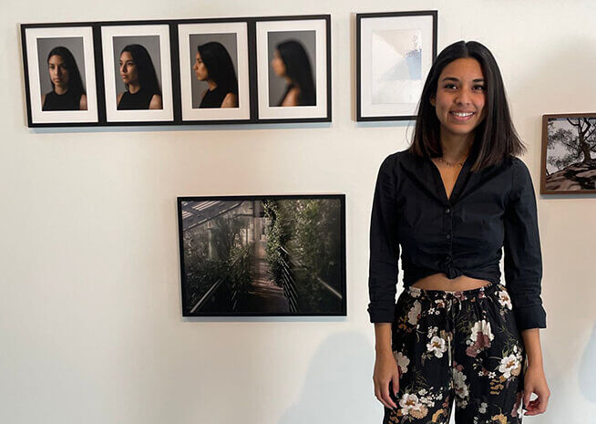 Una dona somrient amb roba casual es troba al costat d'una sèrie de retrats emmarcats i una fotografia de natura, interactuant amb l'art.