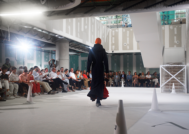 A model walks away from the camera on a fashion show runway, wearing a long black coat with a red hem, creating a striking contrast in front of an attentive audience.