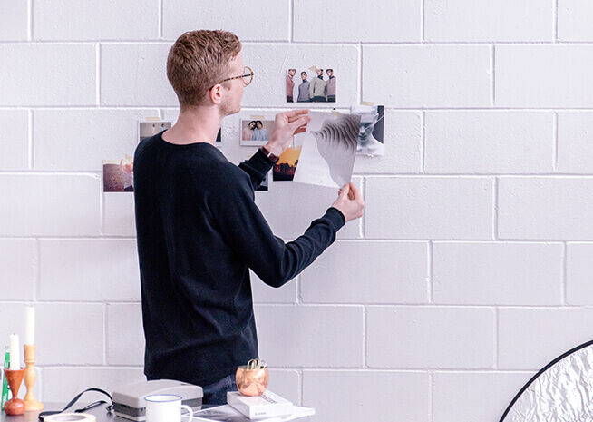 Un hombre con camisa negra ordena una serie de fotos enmarcadas en una pared de ladrillo blanco, creando un espacio de galería personalizado.