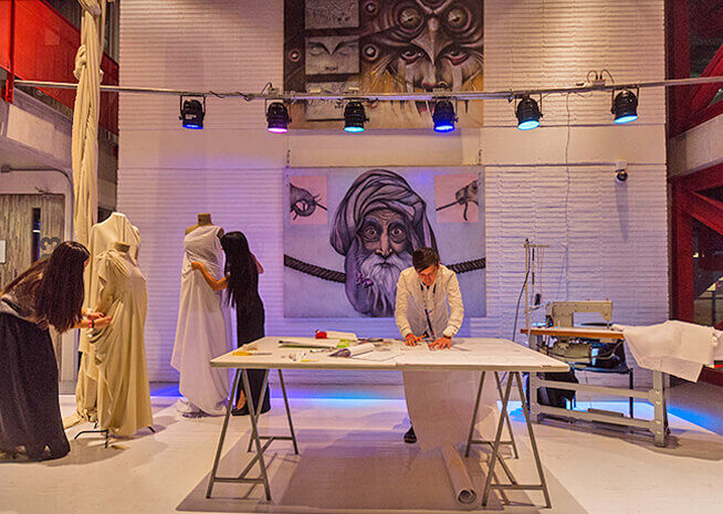 Un artista se enfoca en un dibujo, rodeado de indumentaria cultural y un mural de rostro ampliado en un estudio bien iluminado.