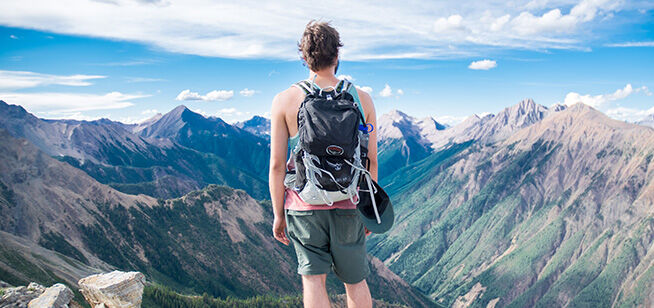 Un hombre con mochila está al borde de una montaña, observando una amplia gama de picos bajo un cielo azul.