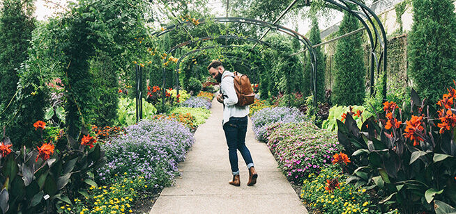 Un hombre contemplativo con mochila camina por un sendero de jardín vibrante, enmarcado por una profusión de flores multicolores y vegetación.