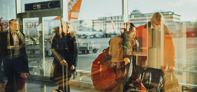 Viajeros con equipaje esperando dentro de una terminal aeroportuaria, bañados en la luz dorada del atardecer, con reflejos y la pista ocupada visible a través de las ventanas.