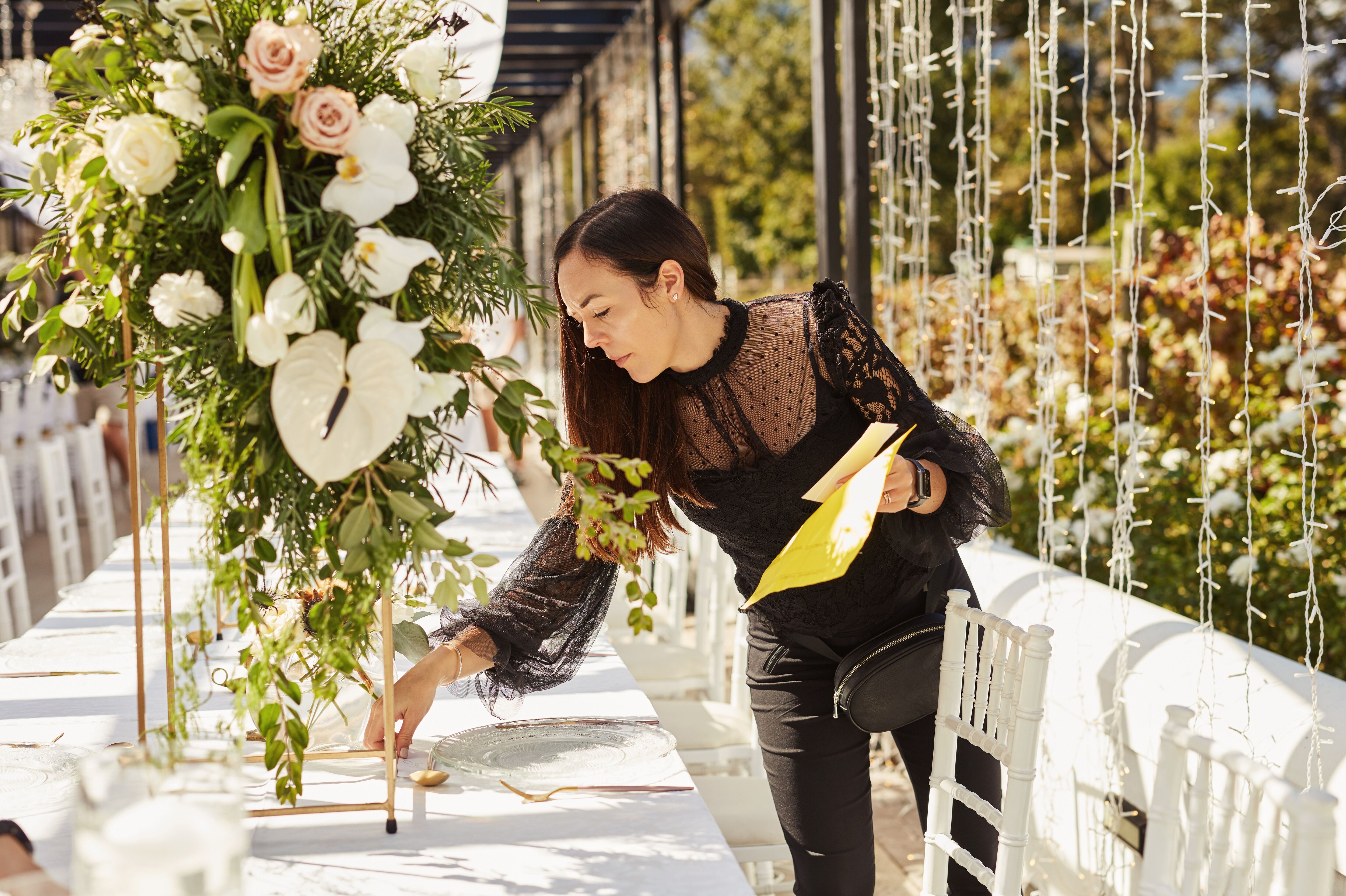 A florist in a black lace blouse is arranging white and pink flowers on a decorated event table, with a focus and attention to detail.