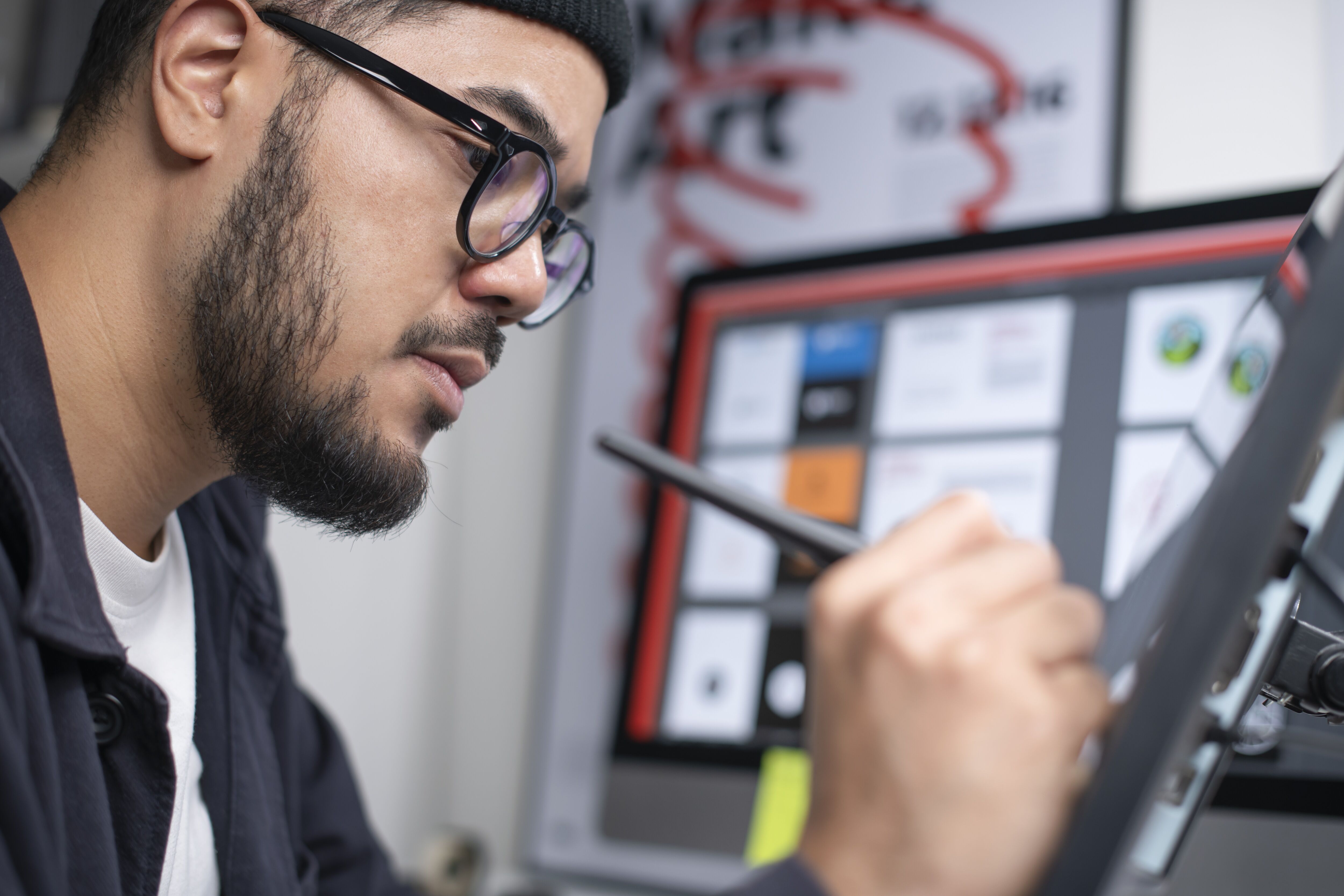 A focused male graphic designer with glasses using a stylus on a digital drawing tablet in a creative workspace.