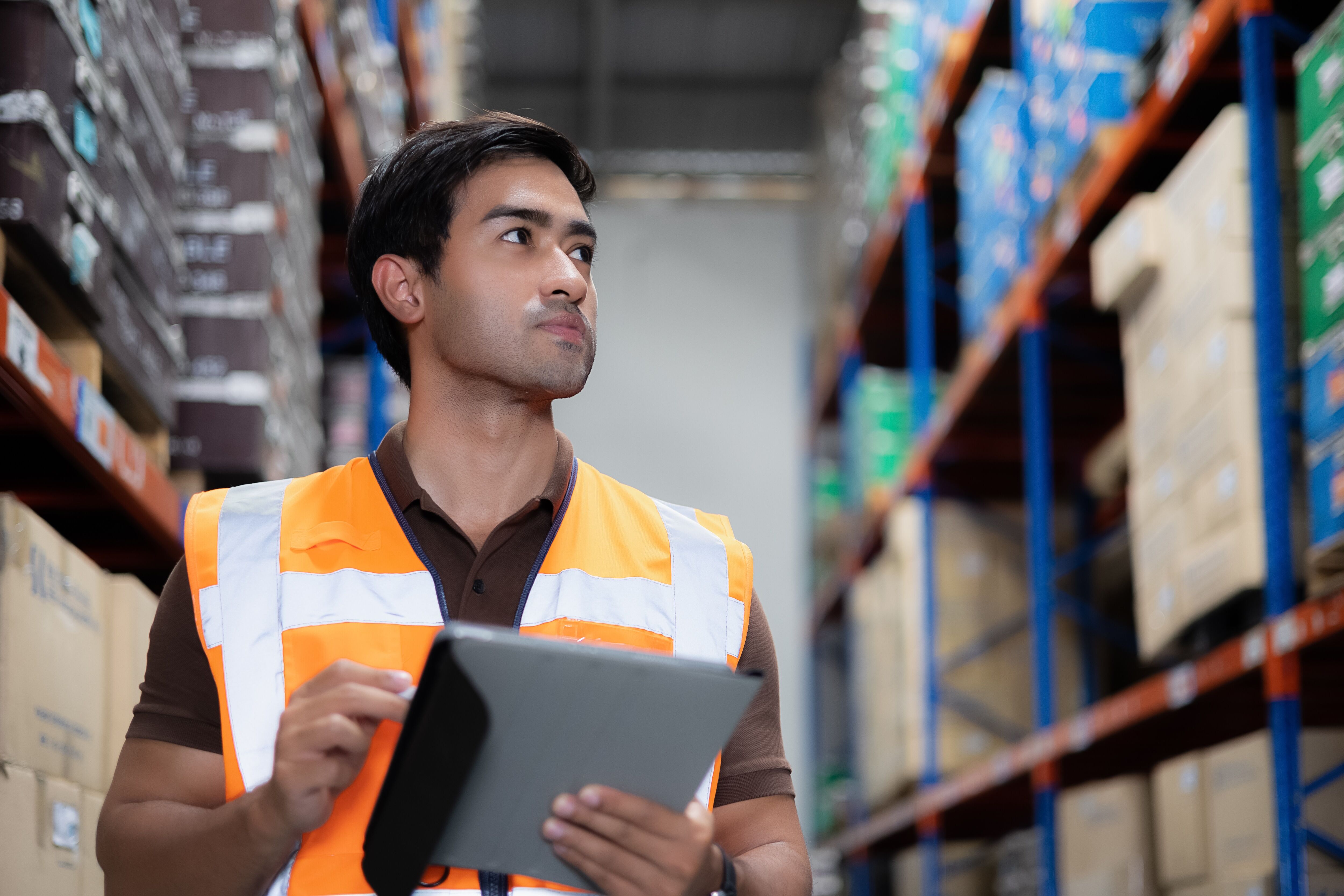 A focused male inventory manager in a high-visibility vest uses a tablet to check stock in a warehouse.