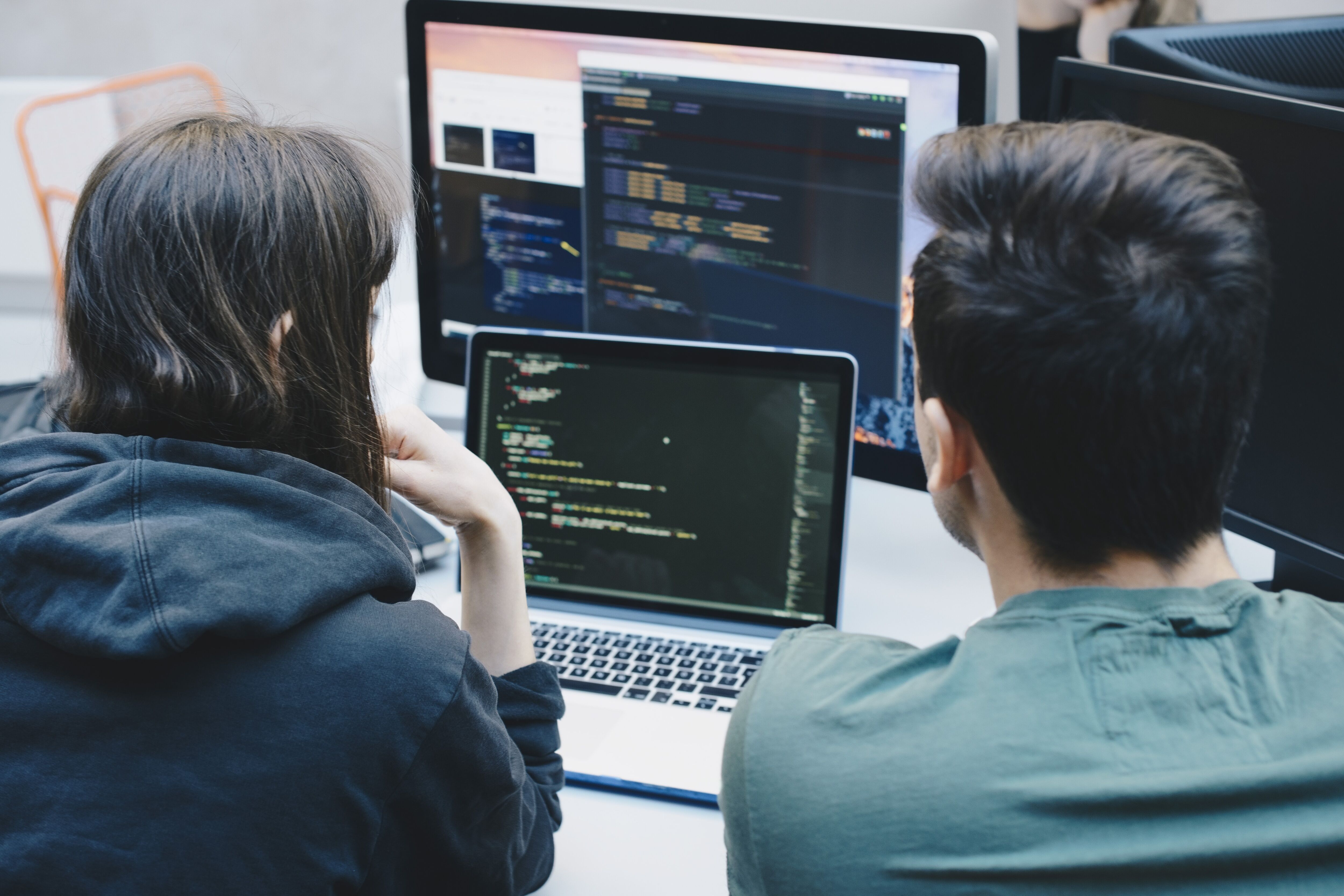 Two professionals code together on a laptop with a dual monitor setup in the background.