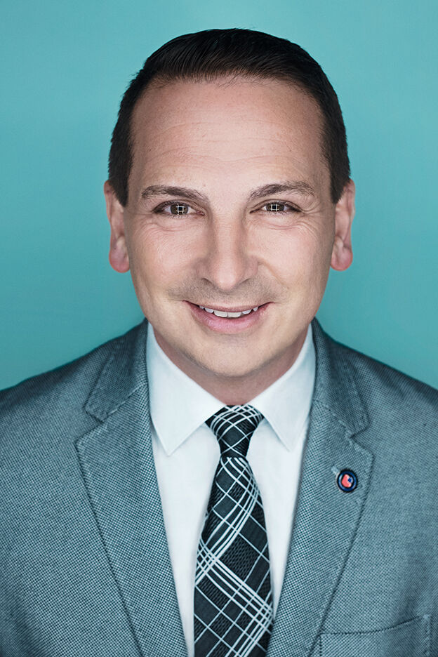 A smiling man wearing a grey suit, white shirt, and plaid tie against a teal background.