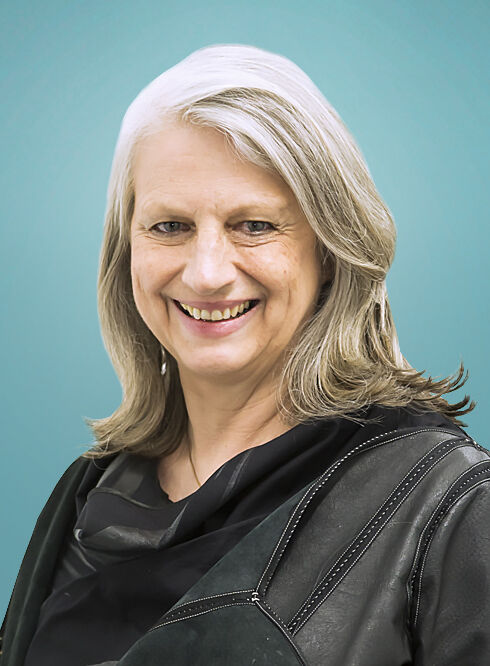 Portrait of a joyful senior woman with shoulder-length silver hair, wearing a black outfit and a leather jacket, set against a turquoise background.