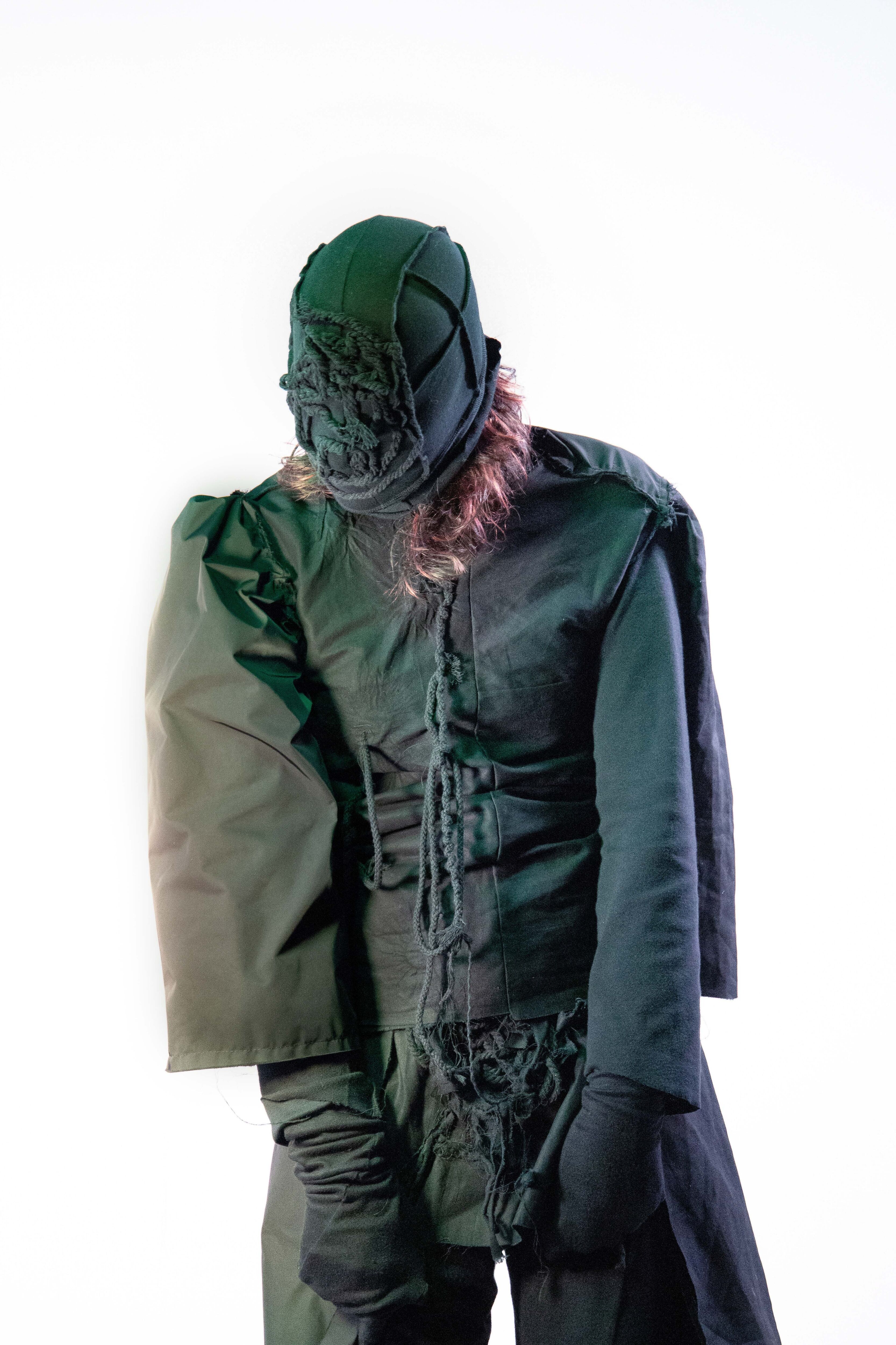 A person shrouded in dark green, textured clothing, with a hood covering their face, stands against a white background, exuding an aura of mystery.
