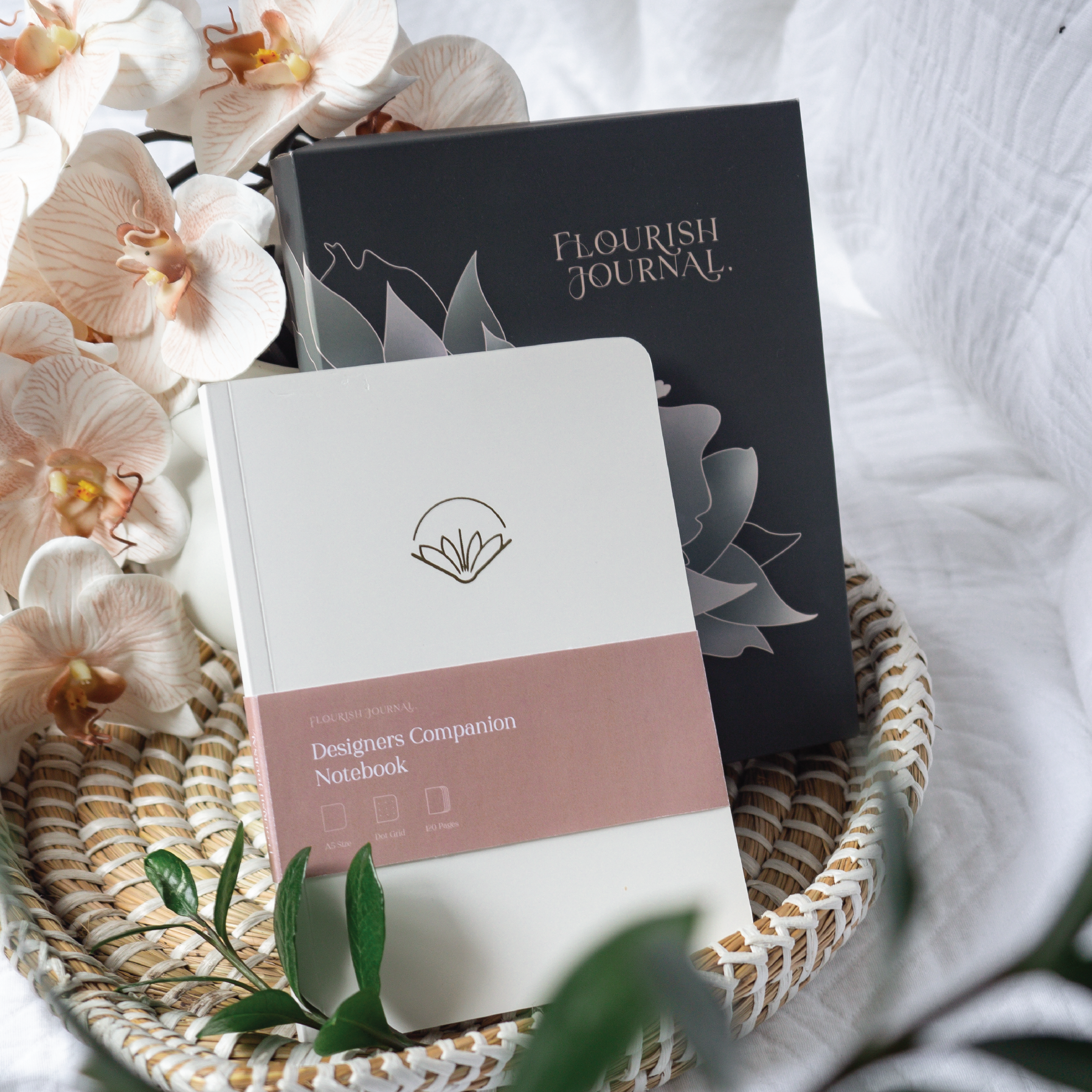 A designer's notebook and journal set, elegantly presented with orchids, in a woven basket on a white cloth background.