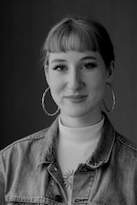A contented young woman with a subtle smile, donning a classic denim jacket and hoop earrings, poses against a gray backdrop.