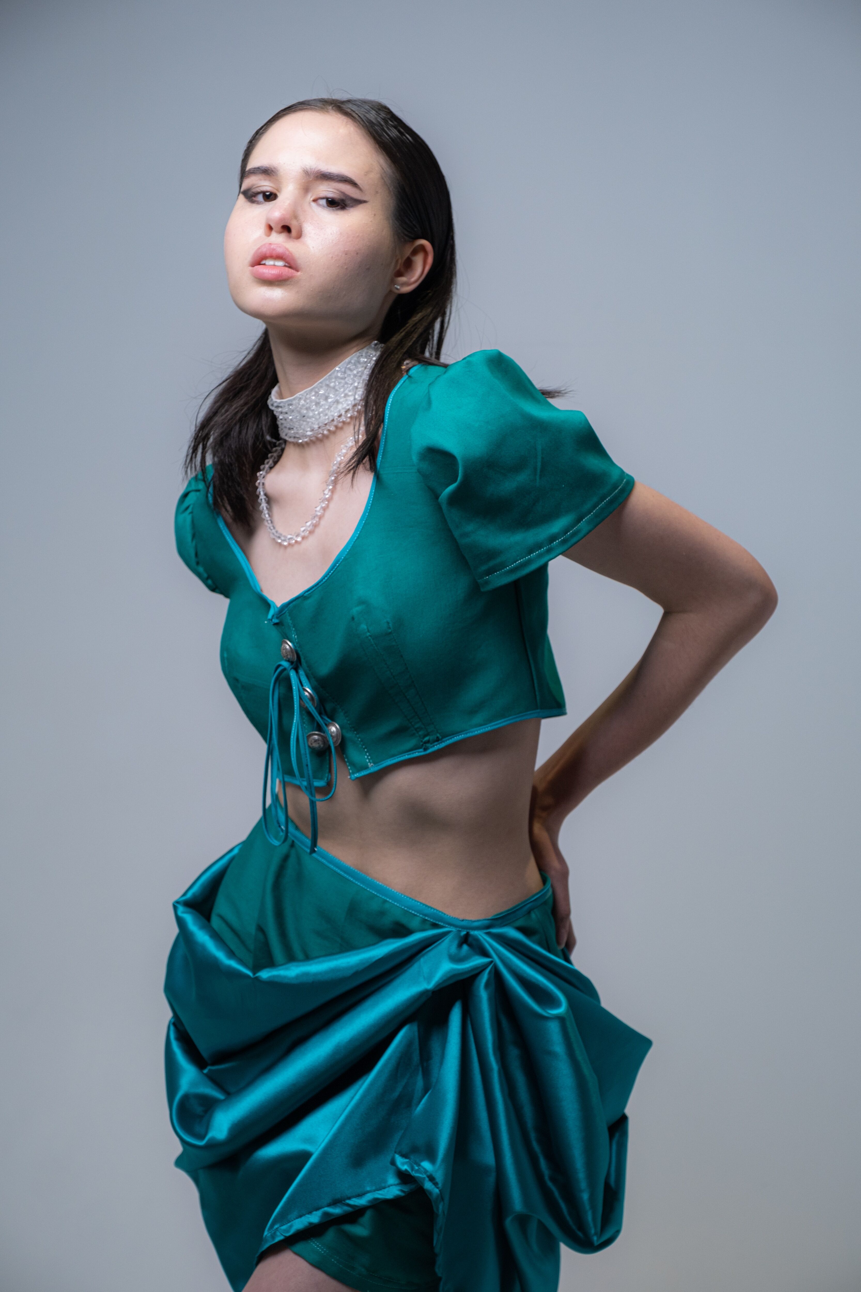  poised model showcasing a modern teal ensemble with a cropped top and draped skirt, accented by a pearl choker.