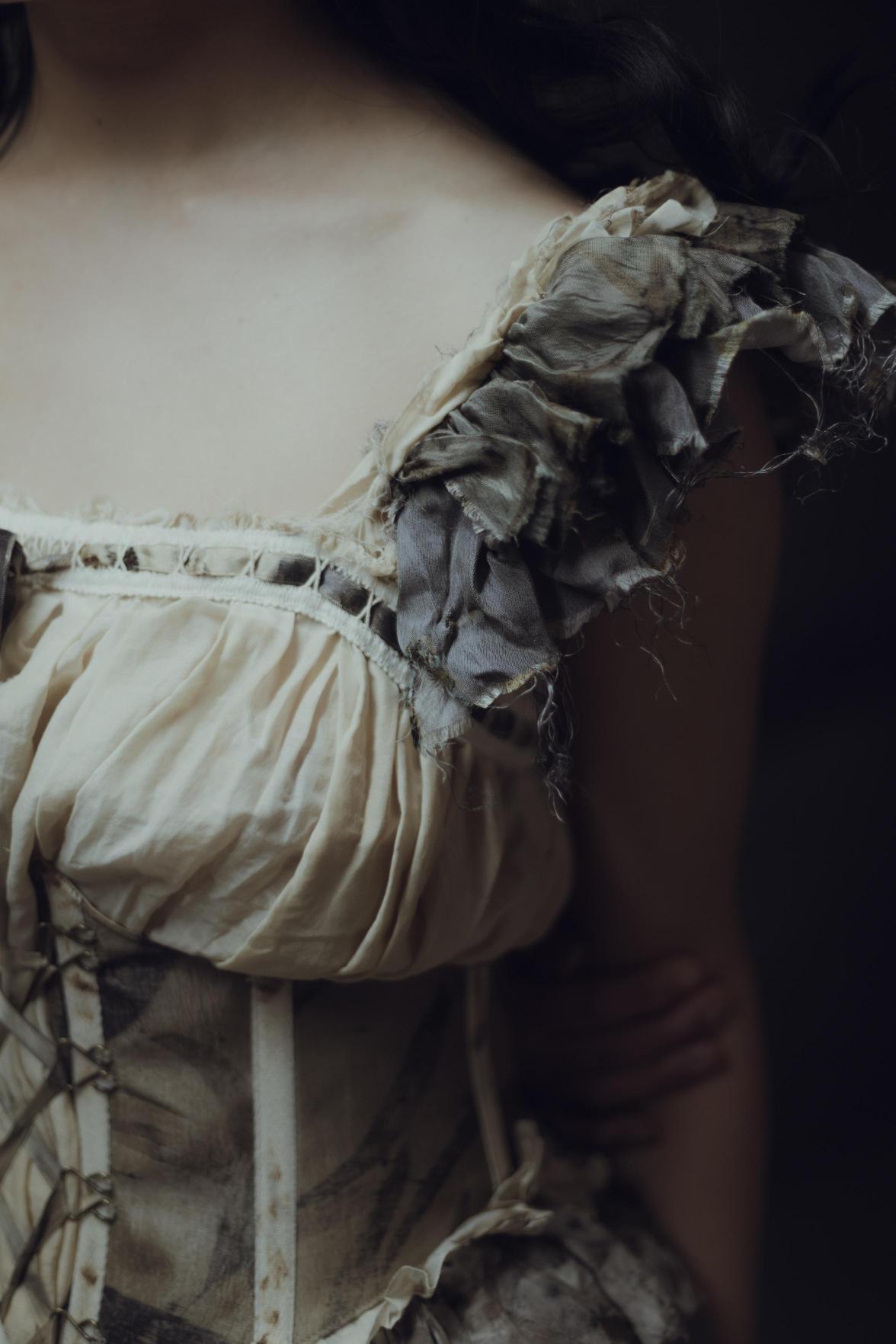 A close-up of a woman's shoulder, wearing a frayed antique dress with intricate lacing.