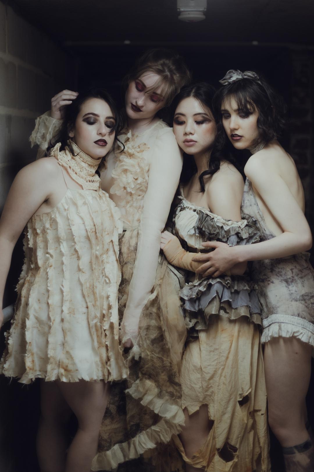Four women in vintage attire pose intimately, their ruffled dresses in muted earth tones complementing the subdued lighting.
