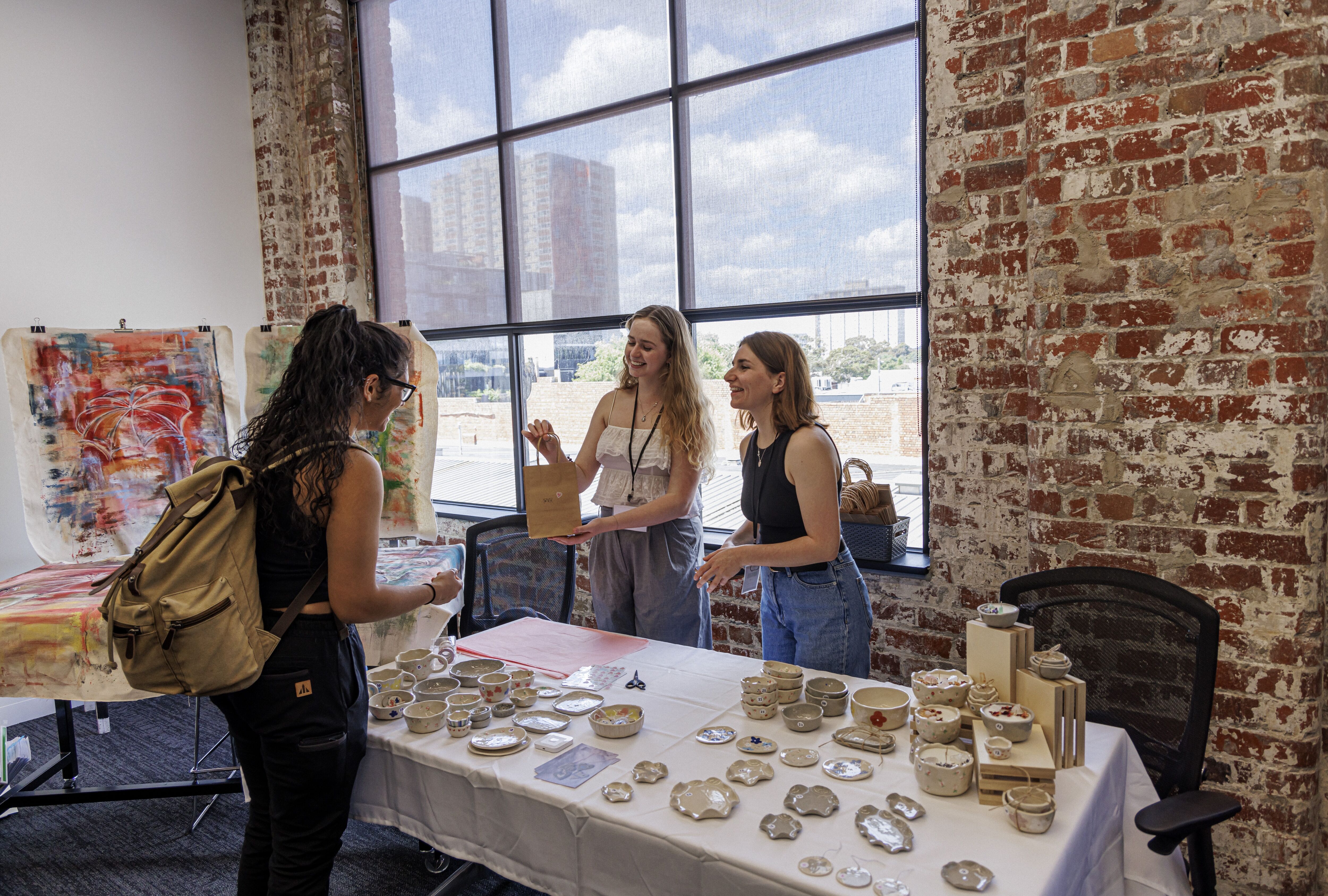 Three women engage at a pottery stall inside a room with large windows and exposed brick, artworks and ceramic wares on display.