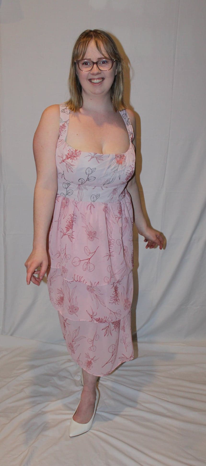 A smiling woman with glasses stands in a sleeveless pink floral dress paired with white shoes.